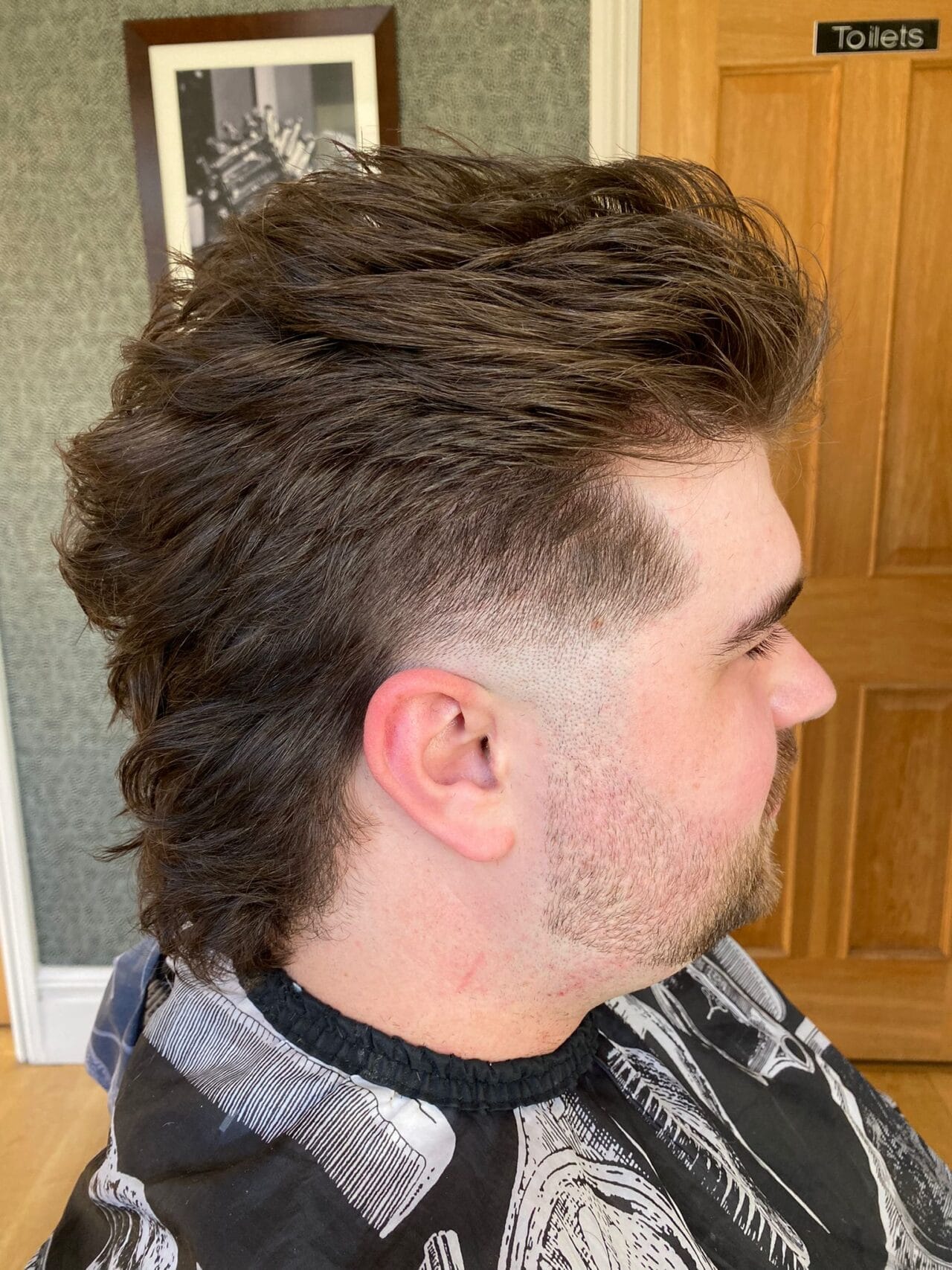 A man with a freshly cut mullet sits in the Barbers Cave chair in Bollington, flaunting short sides and longer, textured hair on top and back. A wooden door labelled "Toilets" and a framed photo are seen in the background.