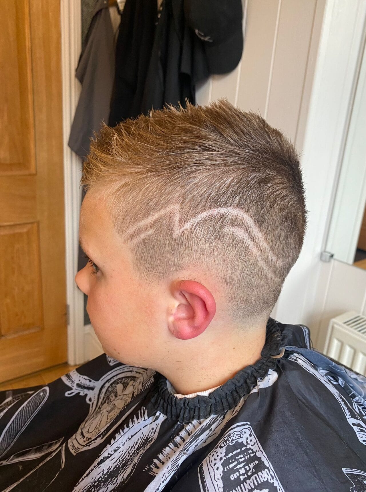 A young boy with short, light brown hair sports a fresh pattern cut by George at The Barber's Cave in Bollington. Shaved zigzag designs adorn the side of his head as he sits in the barbershop, wearing a black haircut gown.